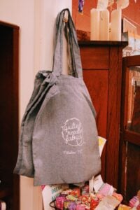 A gray chambray tote bag printed with the Quilted Needle Fabrics logo and "Pittsboro, NC" in white script, hanging on a hook inside the shop with colorful fabric rolls and store merchandise visible below.