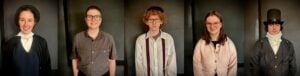 A horizontal row of five individual portraits of young people in period costume, each photographed against a dark gray-green background. From left to right: a girl in a black bonnet, white blouse with high collar, and dark blue vest over a black jacket; a boy with short dark hair and glasses wearing a gray button-up work shirt; a boy with curly red hair, glasses, and a flat cap wearing a white shirt with red and navy striped suspenders; a girl with long brown hair and glasses wearing a soft pink cardigan with buttons over a dark top; and a boy in a tall black top hat and high-collared white cravat under a dark gray frock coat. The styling suggests 19th-century American dress, consistent with a living history program.