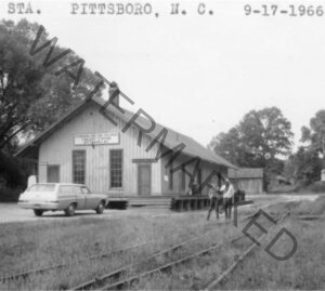A black-and-white photograph labeled "STA. PITTSBORO, N.C. 9-17-1966" across the top. The image shows a long, single-story wooden freight depot with vertical board siding, a pitched roof, and a brick chimney. A sign on the building's near gable end reads "Seaboard Air Line Railway Freight Station, Pittsboro, N.C." Two men stand together on the dirt area in front of the loading platform, and a station wagon is parked at the building's left end. Rusted railroad tracks run through tall grass in the foreground, with mature trees framing the depot on both sides under an overcast sky.