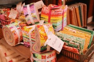 A display of colorful Free Spirit fabric bundles on a wooden table at Quilted Needle Fabrics in Pittsboro, including rolled jelly rolls and stacked fat quarter bundles in vibrant floral, geometric, and tropical prints in shades of yellow, red, green, and pink.