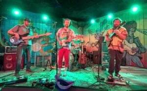 Awen Family Band performing live at The Plant in Pittsboro, bathed in green stage lighting. Three musicians play at the front — a bassist on the left in a cap, a center guitarist in a patterned tee and red pants, and a guitarist on the right in an earth-toned patterned shirt — with a drummer visible behind them. A colorful hand-painted mural covers the back wall, and a floor crowded with pedalboards and cables frames the foreground.