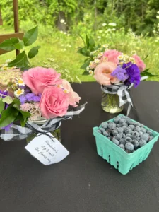 Two mason jar bouquets and a pint of fresh blueberries arranged on a black tablecloth at an outdoor farm stand, with a sunlit meadow of tall grass, wildflowers, and trees in the background. Each jar holds a small mixed bouquet of pink roses and lisianthus, white chamomile daisies, purple ageratum, pink yarrow, and Queen Anne's lace, with leafy greenery and tied with a black and white buffalo-check ribbon. The closer bouquet has a paper gift tag in cursive script that reads "Remember to stop & smell the flowers today" with a small bee illustration. To the right sits a teal-green pulp pint basket overflowing with fresh blueberries dusted with their natural silvery bloom.