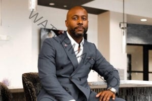 A man in a charcoal double-breasted suit with a white dress shirt, pocket square, and small pink lapel pin, seated in a modern interior setting with neutral tones and pendant lighting.