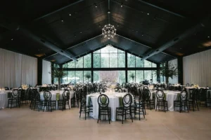 Wedding reception set up inside a modern pavilion with a peaked black wood ceiling, floor-to-ceiling windows overlooking trees, and white curtain-draped walls. Round tables with white linens, sage green napkins, tall fern centerpieces, and black oval-back chairs fill the space beneath a geometric pendant chandelier.