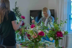 Two women arrange flowers at a long table inside a sunlit room with white walls and a tall window letting in natural light. In the foreground, three glass vases hold loose, garden-style bouquets featuring hot pink peonies, white snapdragons, peach stock, green bells of Ireland, and trailing greenery, with stems and trimmings scattered across the table. On the left, a woman with long blonde hair, wearing a black t-shirt and with a floral tattoo visible on her forearm, is seen from behind as she works on an arrangement. Across the table, a woman with a blonde bob cut, wearing a gray cardigan over a white top, holds a sprig of white flowers and looks down at her bouquet in concentration. The setting suggests an intimate hands-on floral design workshop.