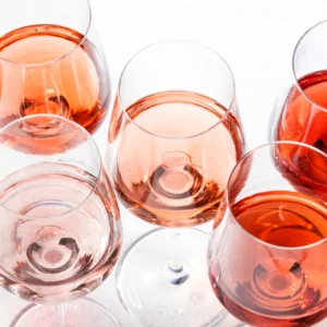 Six stemmed wine glasses filled with varying shades of rosé wine, ranging from pale blush pink to deep amber-rose, arranged on a bright white surface.