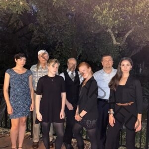 Group portrait of The Lids, a seven-member ensemble posed outdoors at night in front of a wrought-iron railing with trees in the background. The group is dressed in predominantly dark attire, with one person in a patterned vest and tie and another in a blue-patterned dress.