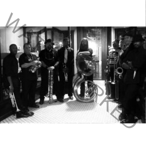 Black-and-white group portrait of a nine-piece brass band standing on a tiled entryway floor, all dressed in black. Instruments include trumpets, a saxophone, a sousaphone, and a trombone. The setting and instrumentation suggest a New Orleans–style brass band.