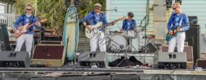 Four-piece band performing on an outdoor stage, all wearing matching blue argyle sweater vests and white pants. Two guitarists flank a bassist and a drummer, with a white surfboard propped upright as a stage prop. A palm tree and pale blue beach house are visible in the background, and a row of monitors and amplifiers lines the stage front. The styling reads as surf rock.