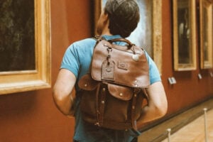 A visitor seen from behind wearing a blue t-shirt and a brown leather backpack, standing in a gallery corridor lined with gold-framed paintings on warm red walls.