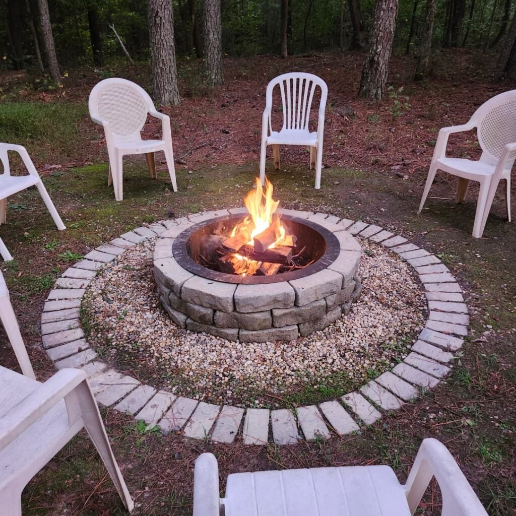 A circular stone firepit at Sky Pond, freshly lit with crackling flames and stacked logs inside a metal liner. The fire pit sits on a tidy gravel-and-paver base in a wooded clearing, encircled by simple white plastic chairs spaced for conversation. Tall pines and hardwoods rise behind, with a soft pine-needle floor in the background.