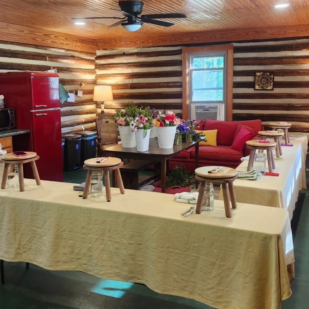 The interior of Sky Pond's tobacco barn cabin, set up for a flower-arranging workshop. Long tables draped in cream linen hold wooden stools, mason jars, and pruning shears at each station; in the center, white buckets brim with fresh tulips, anemones, and ranunculus. A vintage red Galanz refrigerator anchors the kitchen area to the left, with a red couch and yellow accent pillow in the background. The space has log walls, a tongue-and-groove wood ceiling, recessed lighting, and a black ceiling fan.