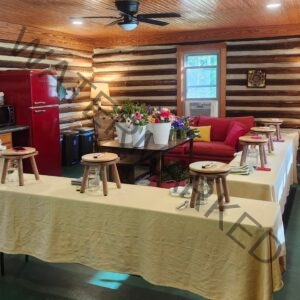 The interior of Sky Pond's tobacco barn cabin, set up for a flower-arranging workshop. Long tables draped in cream linen hold wooden stools, mason jars, and pruning shears at each station; in the center, white buckets brim with fresh tulips, anemones, and ranunculus. A vintage red Galanz refrigerator anchors the kitchen area to the left, with a red couch and yellow accent pillow in the background. The space has log walls, a tongue-and-groove wood ceiling, recessed lighting, and a black ceiling fan.