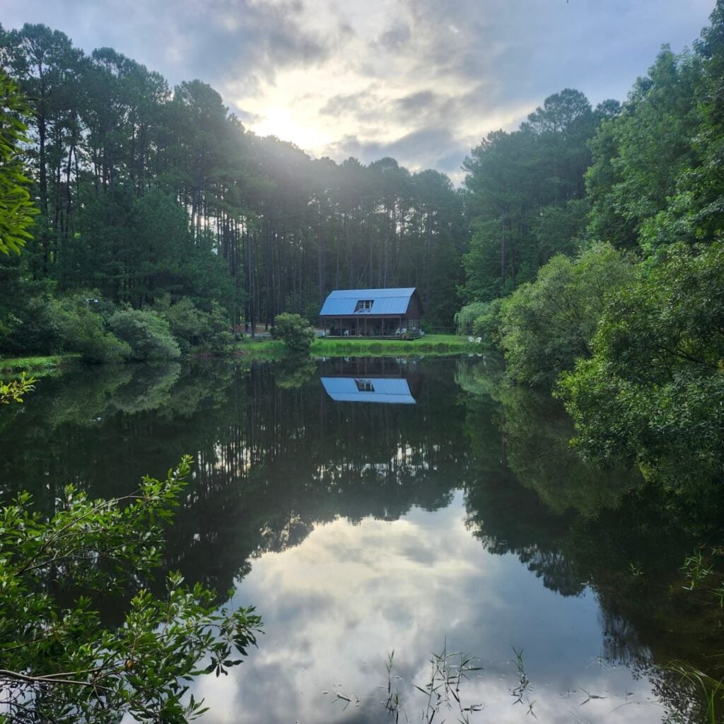 A tranquil view of Sky Pond's renovated tobacco barn cabin, framed across the property's still pond. The two-story cabin with a silver metal roof sits among tall pines on the far shore, with the sun breaking softly through layered clouds above. The pond's glassy surface mirrors the cabin, trees, and dramatic sky almost perfectly, with green foliage framing the foreground.