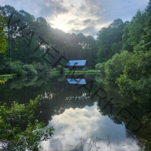 A tranquil view of Sky Pond's renovated tobacco barn cabin, framed across the property's still pond. The two-story cabin with a silver metal roof sits among tall pines on the far shore, with the sun breaking softly through layered clouds above. The pond's glassy surface mirrors the cabin, trees, and dramatic sky almost perfectly, with green foliage framing the foreground.
