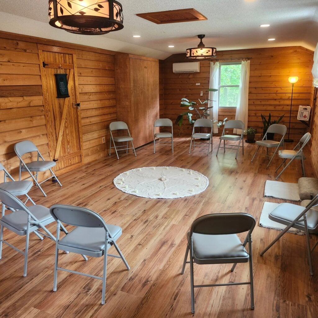 Sky Pond's upstairs studio space at the cabin, set up for a seated group session. Grey padded folding chairs are arranged in a wide circle around a small round white rug holding tealights at its center. The room has warm wood-paneled walls, light hardwood floors, two leafy lantern-style ceiling lights, a window letting in soft daylight, and a few houseplants tucked into the corners.