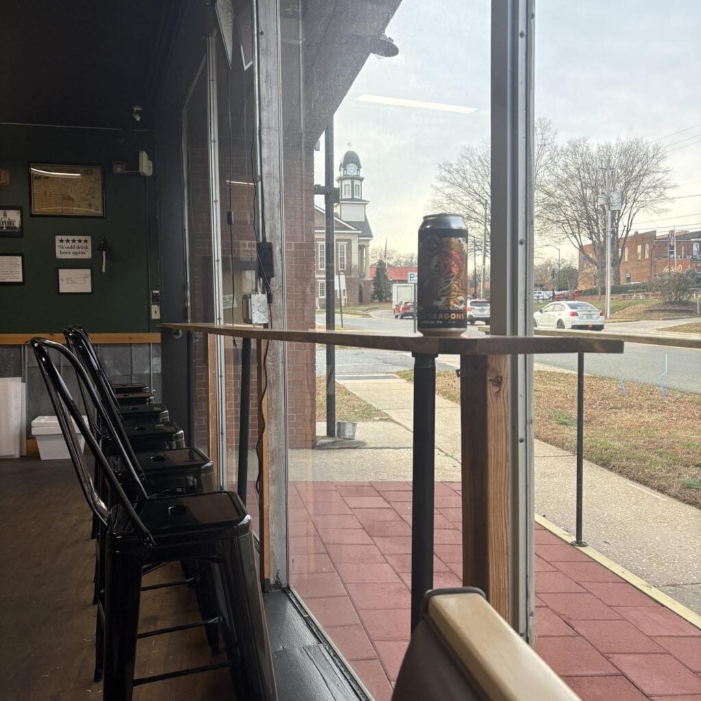 Looking out from the window-side counter at The Place to Be, where a tall can of "Lost Dragons" Imperial IPA sits on the rail. Through the glass, Pittsboro's historic Chatham County Courthouse with its white clock tower rises across the street, with downtown brick storefronts and parked cars in view. Inside the bar, black metal stools line the counter, and a framed five-star review reading "Would drink here again" hangs on the green wall behind.