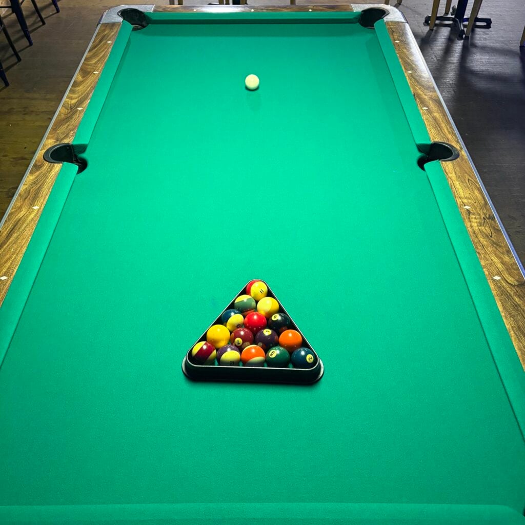 Top-down view of The Place to Be's pool table, with bright green felt and dark wood rails. All 15 numbered balls are racked tightly in the black plastic triangle near the foot of the table, and the white cue ball sits placed for the break. Cue sticks and chairs are visible at the edges of the frame.