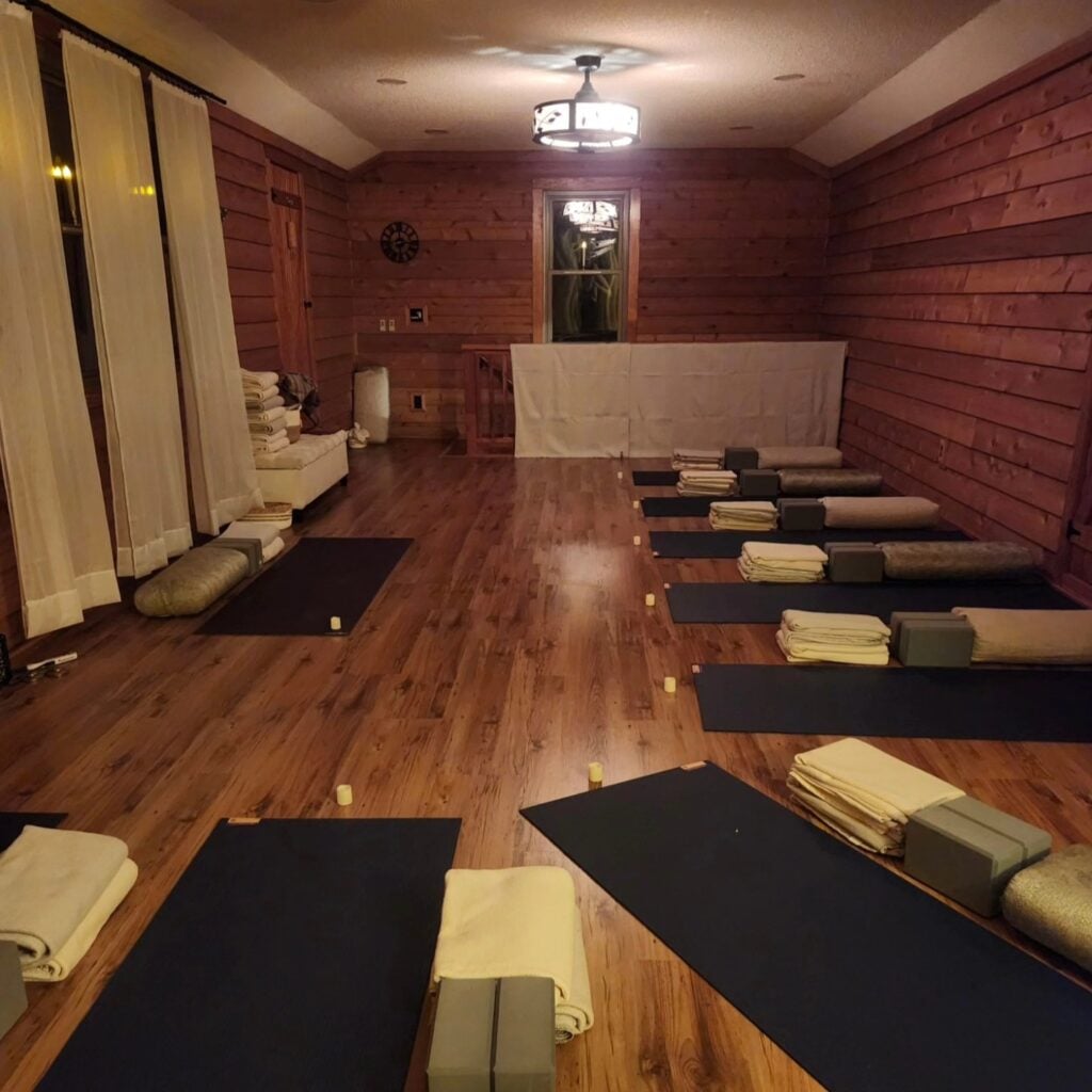 Sky Pond's upstairs studio set up for a candlelit yoga class. Two rows of black yoga mats line the warm-toned hardwood floor, each paired with a neatly folded cream blanket, a sage block, and a linen bolster. Small electric candles are placed between mats. The room has warm cedar-toned wood-paneled walls, soft curtains framing the windows, and a Mission-style ceiling lamp casting golden light overhead.