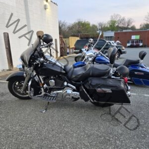 A row of Harley-Davidson cruisers parked side by side outside The Place to Be on a warm evening. A black bike with a windshield and saddlebags sits in the foreground, with two blue baggers parked behind it. The bar's white side wall and lattice patio fencing frame the lot, with downtown Pittsboro buildings visible in the background.