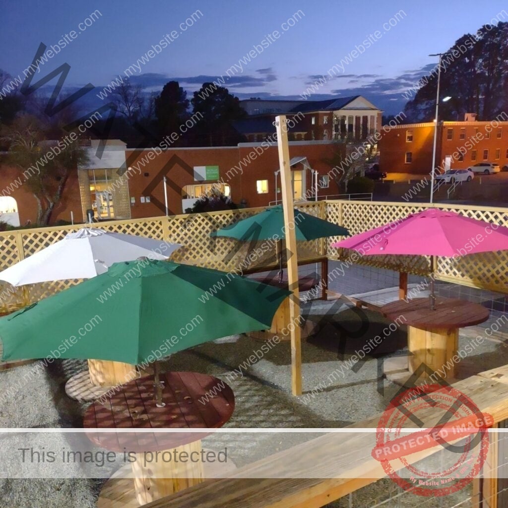 Twilight view of The Place to Be's outdoor patio, with repurposed wooden cable-spool tables shaded by colorful market umbrellas in green, pink, and white. The space is enclosed by a tan wood lattice fence and finished in gravel underfoot. Beyond the patio, the lit windows of downtown Pittsboro's brick buildings glow against a deep blue evening sky.