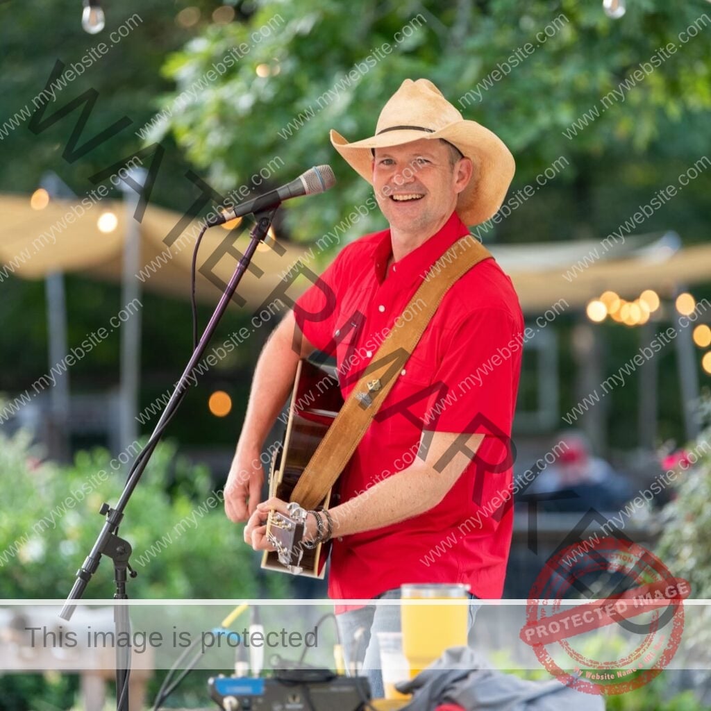 A musician performs at an outdoor evening event at Old Mill Farm, smiling toward the audience. He wears a straw cowboy hat, a bright red short-sleeved snap shirt, and jeans, and plays an acoustic guitar with a leather strap. Behind him, sail shades and warm string lights glow above the venue's open seating area at golden hour.