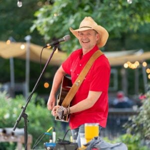 A musician performs at an outdoor evening event at Old Mill Farm, smiling toward the audience. He wears a straw cowboy hat, a bright red short-sleeved snap shirt, and jeans, and plays an acoustic guitar with a leather strap. Behind him, sail shades and warm string lights glow above the venue's open seating area at golden hour.