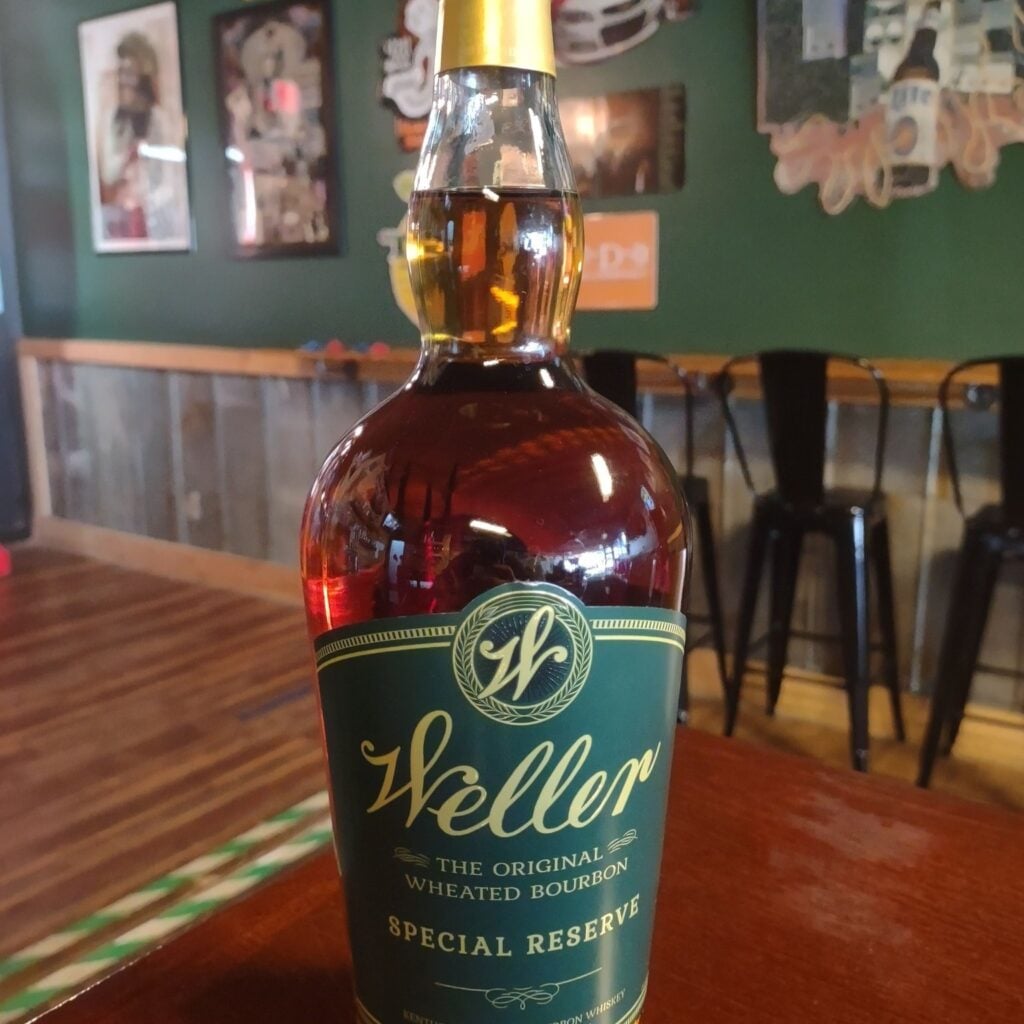 A bottle of W.L. Weller Special Reserve wheated bourbon, with its distinctive green and gold label, photographed on a wooden tabletop inside The Place to Be. The bar's deep green walls hold framed band posters and music memorabilia, with black metal bar stools and a wood counter softly blurred behind.