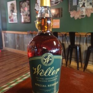 A bottle of W.L. Weller Special Reserve wheated bourbon, with its distinctive green and gold label, photographed on a wooden tabletop inside The Place to Be. The bar's deep green walls hold framed band posters and music memorabilia, with black metal bar stools and a wood counter softly blurred behind.
