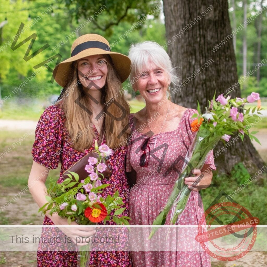 Two women stand side by side under shade trees at Old Mill Farm, both smiling at the camera and holding bouquets of fresh-cut flowers including pink campanula, lilies, and orange poppies. The younger woman on the left wears a wide-brimmed straw sun hat and a deep pink floral dress; the older woman on the right has white hair and wears a soft pink floral dress with sunglasses tucked into the neckline.