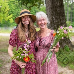 Two women stand side by side under shade trees at Old Mill Farm, both smiling at the camera and holding bouquets of fresh-cut flowers including pink campanula, lilies, and orange poppies. The younger woman on the left wears a wide-brimmed straw sun hat and a deep pink floral dress; the older woman on the right has white hair and wears a soft pink floral dress with sunglasses tucked into the neckline.