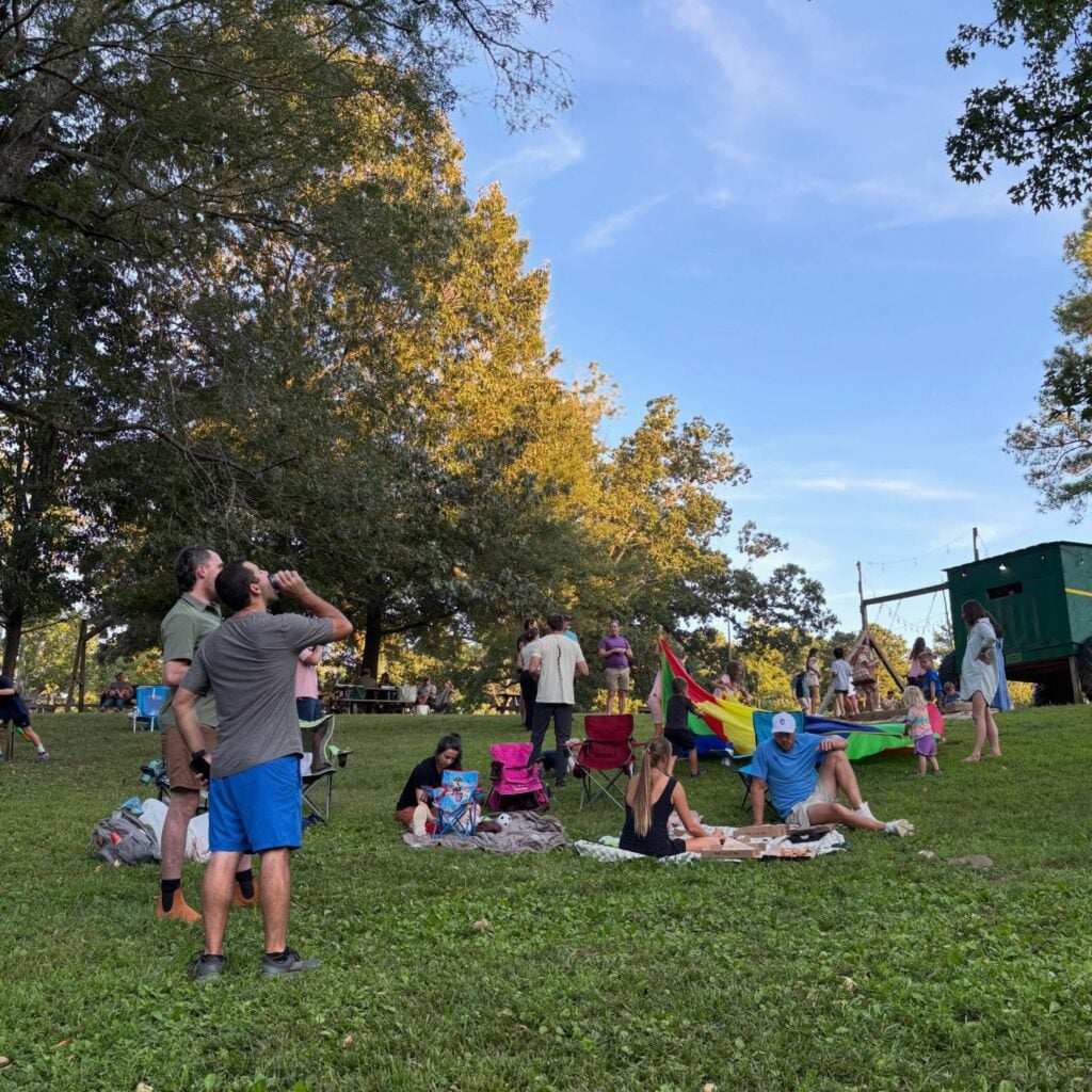 A wide view of a sunlit lawn at Old Mill Farm during an evening event. Adults stand or sit in folding camp chairs and on picnic blankets — some sipping drinks, others sharing pizza — while children play with a large rainbow parachute toward the back of the frame. A wooden treehouse-style structure with string lights sits at the right edge, and tall oaks frame a soft blue sky above.