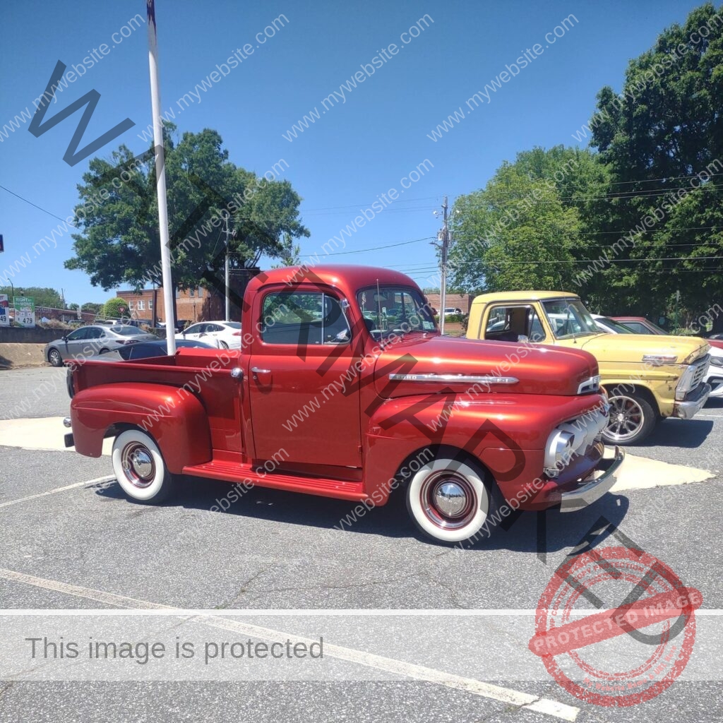 A glossy restored mid-century Ford F-1 pickup truck in candy-apple red, parked in The Place to Be's lot under a clear blue sky. Its whitewall tires and polished chrome bumper catch the sun. A yellow vintage Ford pickup is parked just behind it, with downtown Pittsboro storefronts and a flagpole visible in the background.