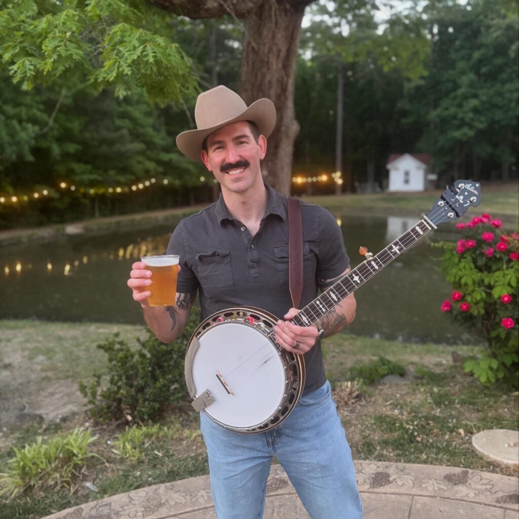 A musician stands on a stone patio beside Old Mill Farm's pond at dusk, smiling at the camera. He wears a tan cowboy hat, a dark short-sleeved henley, and jeans, and holds a Gibson banjo across his body with one hand and a plastic cup of golden beer in the other. String lights twinkle along the pond's edge behind him, and a red rosebush blooms to the right.