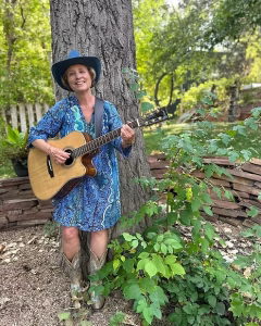 A musician poses outdoors against the trunk of a tall tree on a sunny day, smiling warmly at the camera. She wears a flowing blue paisley dress, a blue cowboy hat, and metallic silver cowboy boots, and holds an acoustic guitar across her body. A low stone wall, leafy green plants, and a softly blurred garden setting fill the background.