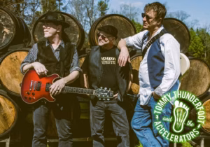 A promotional photo of the band Tommy Thunderfoot and the Accelerators. Three musicians pose in front of a tall stack of wooden whiskey or wine barrels in an outdoor setting. On the left, a man in a black hat, sunglasses, and a black vest holds a sunburst electric guitar. In the center, an older man in a black newsboy cap, sunglasses, and a dark band t-shirt leans casually with his arm on the right musician's shoulder. On the right, a tall man in sunglasses, a blue shirt, and white tee stands with hands in his pockets. The band's circular green-and-cream logo featuring a guitar headstock graphic sits in the bottom right corner.
