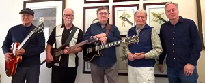 A five-member band poses together for a portrait in front of a gallery wall hung with framed botanical and bird illustrations. From left to right: a man in a black cap and dark sweater holding a sunburst electric guitar; a man with grey hair and a goatee in a black vest with an electric bass; a man with glasses and dark hair in a navy shirt holding a black electric guitar; a man with white hair in a checked shirt and dark vest holding an acoustic guitar; and a man in a dark button-up with hands clasped at his side. All five smile at the camera.