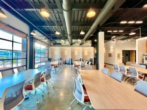 A bright, open interior space with an industrial design aesthetic. The high ceiling is left exposed, painted dark, with visible ductwork, pipes, and conduit, and is lit by a mix of dome-shaped pendant lights and recessed fixtures. Polished concrete floors run throughout. Several long tables with light wood tops are arranged in rows, surrounded by rolling office chairs with white frames, gray mesh backs, and rust-orange seat cushions. Along the back wall, upholstered booth seating sits beneath a series of wall sconces and softly patterned wall panels. Tall, black-framed garage-style windows line the left side, letting in daylight and revealing trees, parked cars, and nearby buildings outside. A white structural column stands at the center, and a seating lounge with framed black-and-white photographs and a coffee station is visible to the right.
