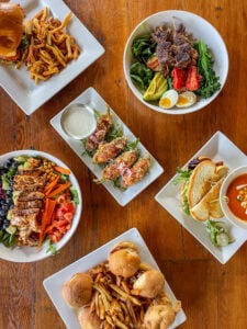 Flat-lay food photography showing six plated dishes arranged on a warm, rustic wooden table. Clockwise from upper left: a burger on a brioche bun with a generous pile of shoestring fries; a grain bowl topped with skewered steak, crispy pork belly, roasted tomatoes, broccolini, avocado, spinach, and a soft-boiled egg; a half-sandwich on toasted bread paired with a cup of creamy tomato soup garnished with mozzarella; pulled pork sliders on soft buns served with seasoned fries; a chopped salad with grilled chicken, blueberries, chickpeas, julienned carrots, diced tomatoes, and pickled red cabbage; and a center plate of breaded fried jalapeño peppers topped with pickled onions and parmesan, served with a ramekin of ranch dressing on a bed of arugula.