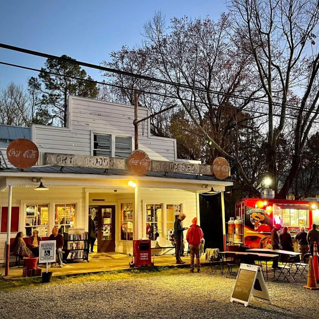 Inside the Bynum General Store, a cloth-covered table holds dozens of vintage drinking glasses printed with old soda logos like Pepsi, Mountain Dew, and Coca-Cola, with more displayed in a wooden bottle crate at left. Tall shelves behind hold antique tins, bottles, and general-store memorabilia, and a man in a vest and knit cap works behind the counter at right.