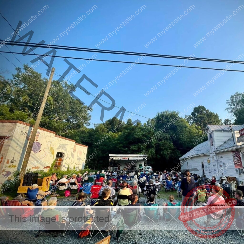 A large audience sits in folding camp chairs on a gravel and grass lot, facing a small covered stage set up for the Bynum Front Porch music series. The white clapboard general store stands at right with a Coca-Cola sign, and a building painted with a colorful animal mural is at left. Tall green trees and a clear evening sky frame the scene, with power lines crossing overhead.