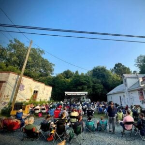 A large audience sits in folding camp chairs on a gravel and grass lot, facing a small covered stage set up for the Bynum Front Porch music series. The white clapboard general store stands at right with a Coca-Cola sign, and a building painted with a colorful animal mural is at left. Tall green trees and a clear evening sky frame the scene, with power lines crossing overhead.