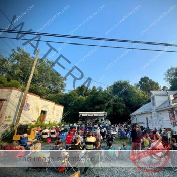 A large audience sits in folding camp chairs on a gravel and grass lot, facing a small covered stage set up for the Bynum Front Porch music series. The white clapboard general store stands at right with a Coca-Cola sign, and a building painted with a colorful animal mural is at left. Tall green trees and a clear evening sky frame the scene, with power lines crossing overhead.
