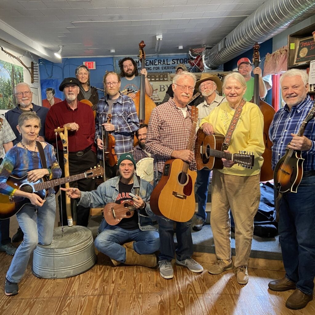 About fifteen musicians of varying ages gather for a group photo inside the Bynum General Store, smiling and holding their instruments — acoustic guitars, a twelve-string, banjos, a fiddle, a mandolin, ukulele, harmonica, and two upright basses. A washtub bass stands in the foreground. Behind them are blue-painted walls with a hand-painted "Bynum General Store" sign, vintage shelving, and exposed ductwork along the ceiling.
