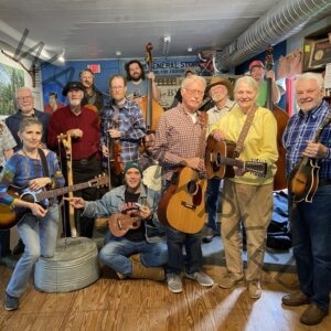 About fifteen musicians of varying ages gather for a group photo inside the Bynum General Store, smiling and holding their instruments — acoustic guitars, a twelve-string, banjos, a fiddle, a mandolin, ukulele, harmonica, and two upright basses. A washtub bass stands in the foreground. Behind them are blue-painted walls with a hand-painted "Bynum General Store" sign, vintage shelving, and exposed ductwork along the ceiling.