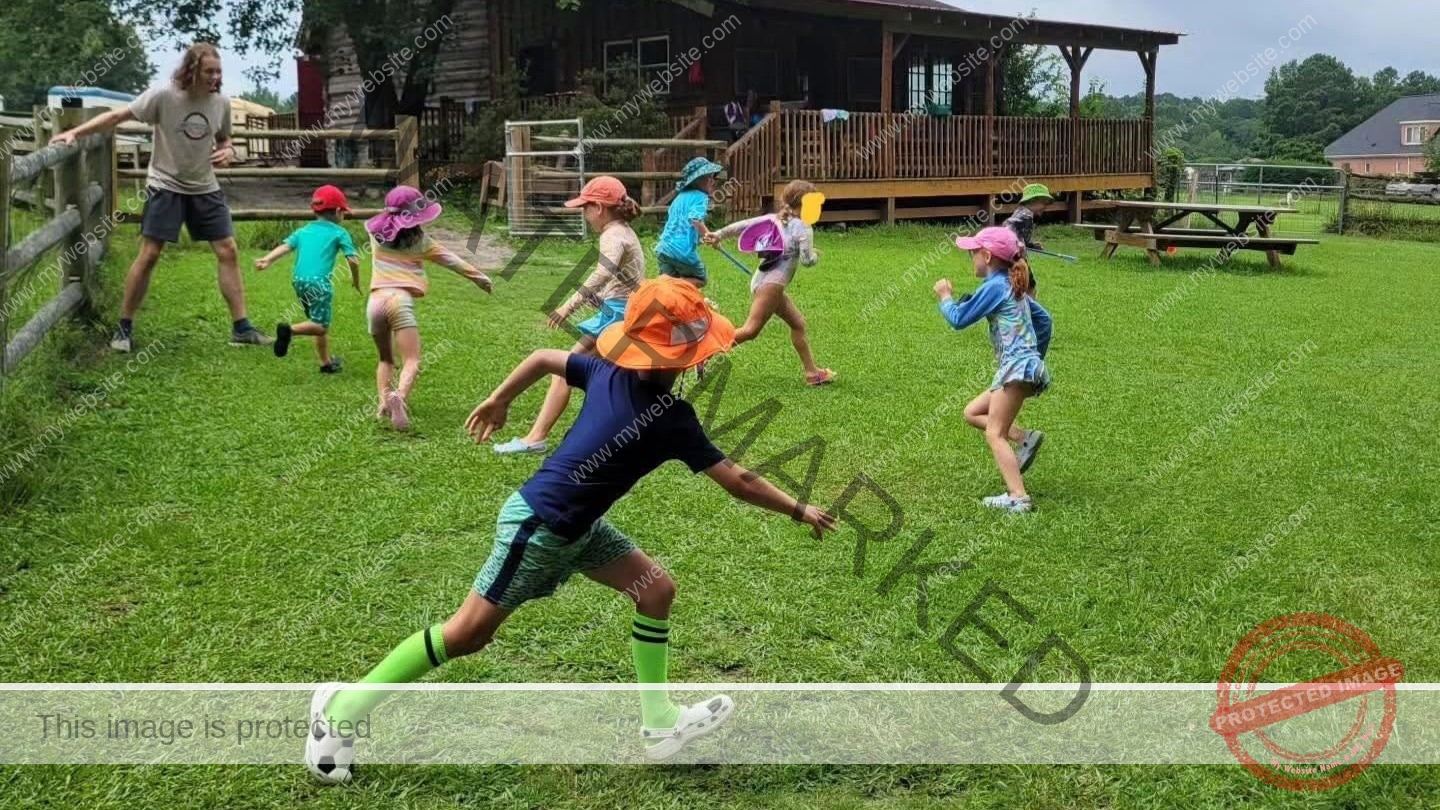 About eight children in colorful sun hats and athletic clothes run and chase across a wide grassy lawn during an outdoor game, one in the foreground sprinting toward a soccer ball. A counselor stands at a wooden fence on the left, and a large rustic timber barn with a covered porch and a picnic table sits in the background under an overcast sky.
