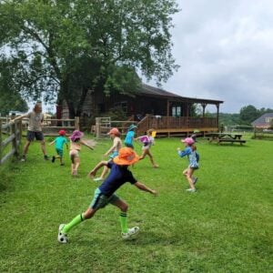 A group of children run and play across a grassy lawn, one in front kicking a soccer ball, while a counselor stands by a wooden fence. A large rustic timber barn with a covered porch and a picnic table sits in the background.