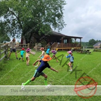 A group of children run and play across a grassy lawn, one in front kicking a soccer ball, while a counselor stands by a wooden fence. A large rustic timber barn with a covered porch and a picnic table sits in the background.
