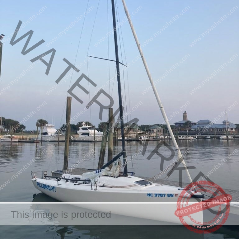A small white racing sailboat with its mast up and sails stowed sits tied to a wooden dock in calm water. Behind it, a marina of larger powerboats and yachts, palm trees, and low waterfront buildings stretches under a pale evening sky.