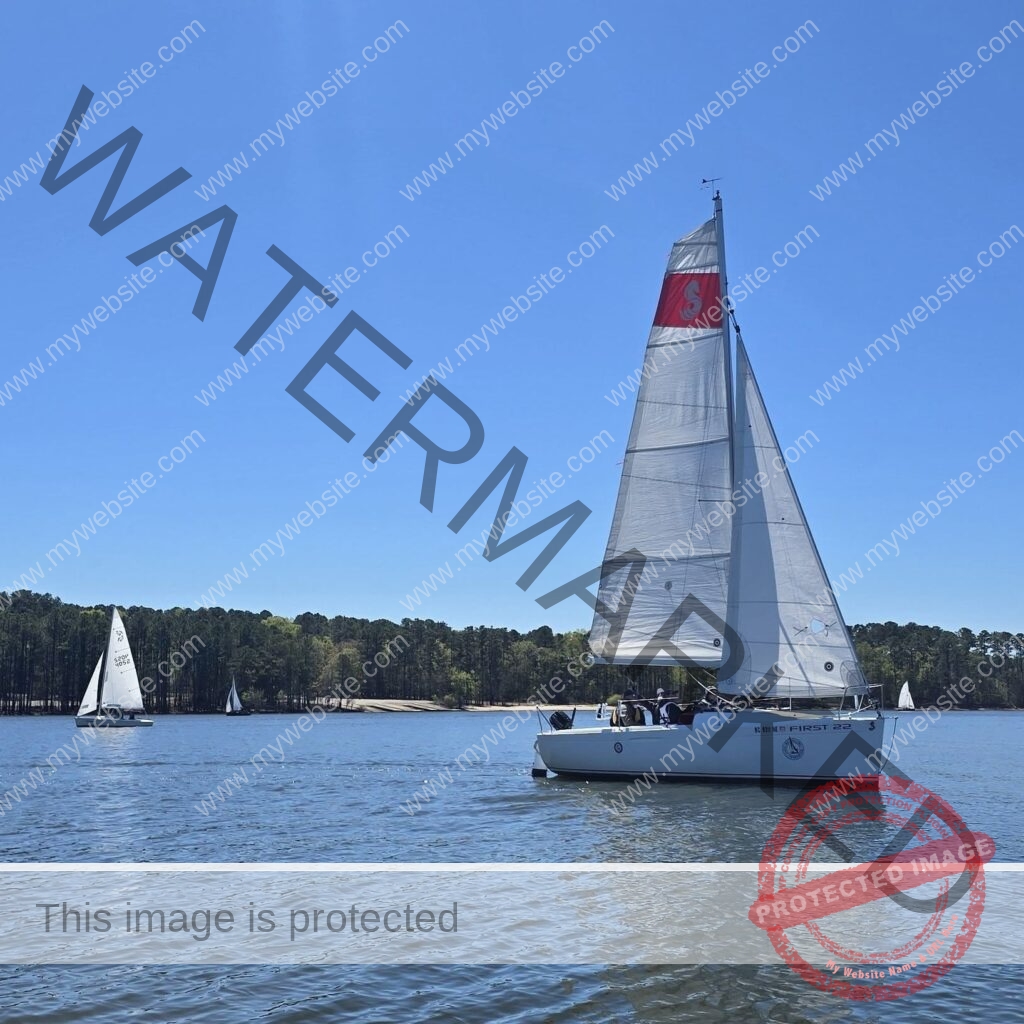 A white keelboat (a Beneteau First 22) sails across open lake water under full sail, its mainsail marked with a red panel and a seahorse-style logo. Two smaller sailboats and a low, wooded shoreline with a sandy bank sit in the background under a clear blue sky.